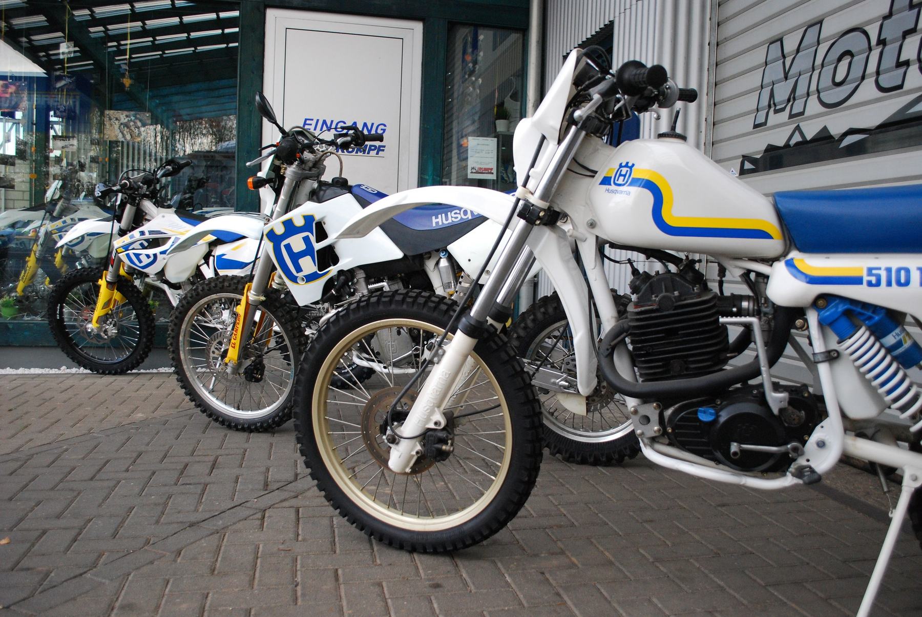 Drei Generationen Husqvarna-Enduros vor dem Eingang Moto Rumpf, vorn die klassische TE 510 in Weiß-Blau-Gelb